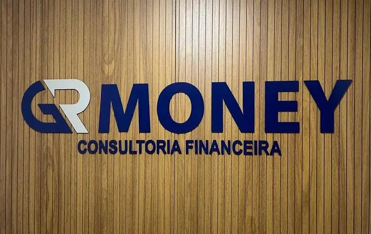 Foto da entrada do escritório da GR Money. Na foto, há um letreiro da logo da empresa escrito em azul colado em uma parede de madeira ripada.